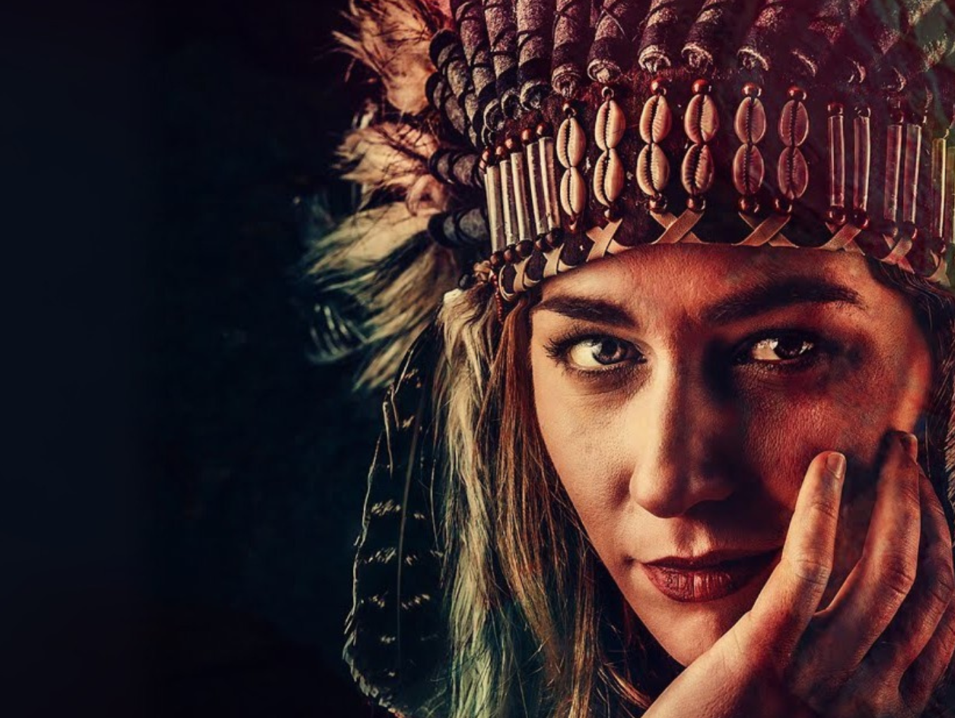 A woman wearing a traditional-style feathered headdress adorned with beads and shells, gazing directly at the camera with her hand resting against her face. The background is dark, highlighting her face and the detailed textures of the headdress.
