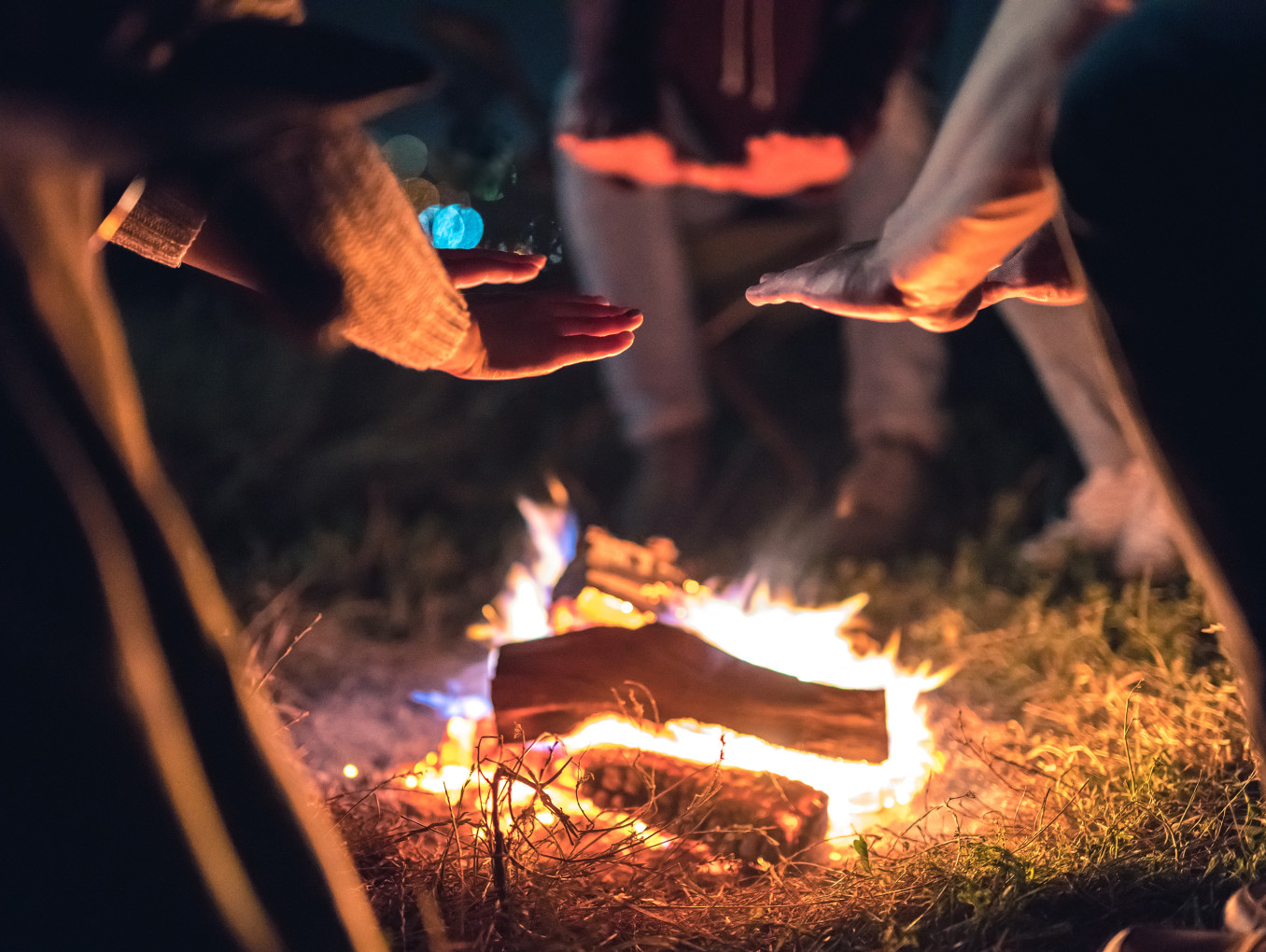 This image captures a cozy outdoor scene where people are gathered around a small campfire at night. Several hands are extended toward the fire, likely to warm up, as the flames crackle over the logs. The soft glow of the fire illuminates the surrounding area, casting warm light on the hands and nearby grass.