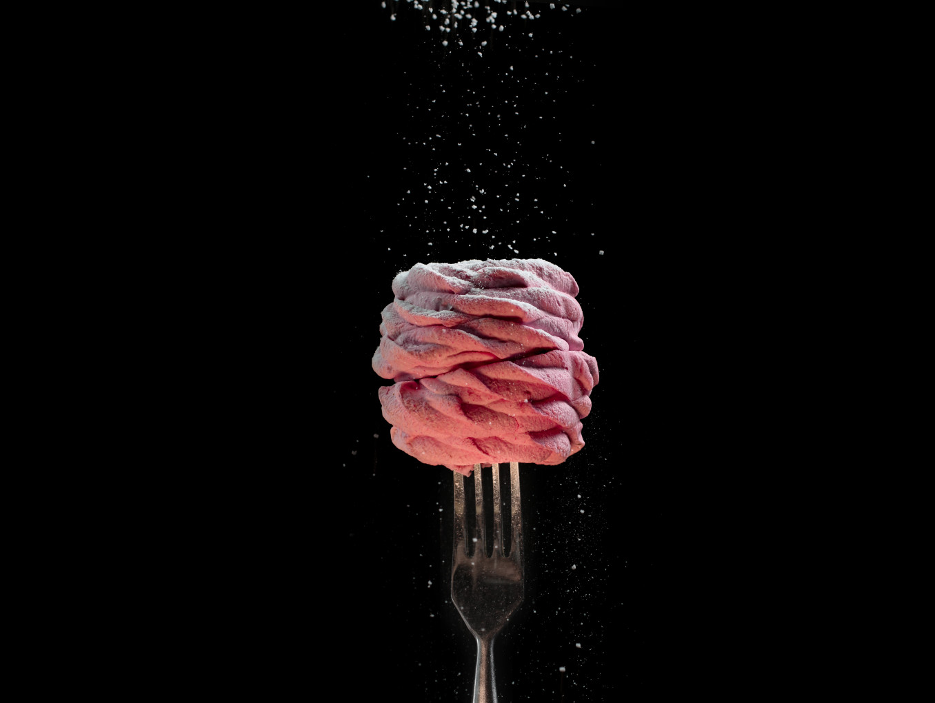 The image depicts a visually striking food item, likely a dessert, against a black background. The dessert consists of twisted pink meringue or a similar pastry, reminding human brain structure, sitting atop a fork that is held upright. Powdered sugar is being sprinkled over the dessert, creating a delicate dusting effect, with fine particles of sugar visible in mid-air. The use of contrasting dark and light elements, as well as the minimalistic setup, gives the image a refined, artistic feel. 