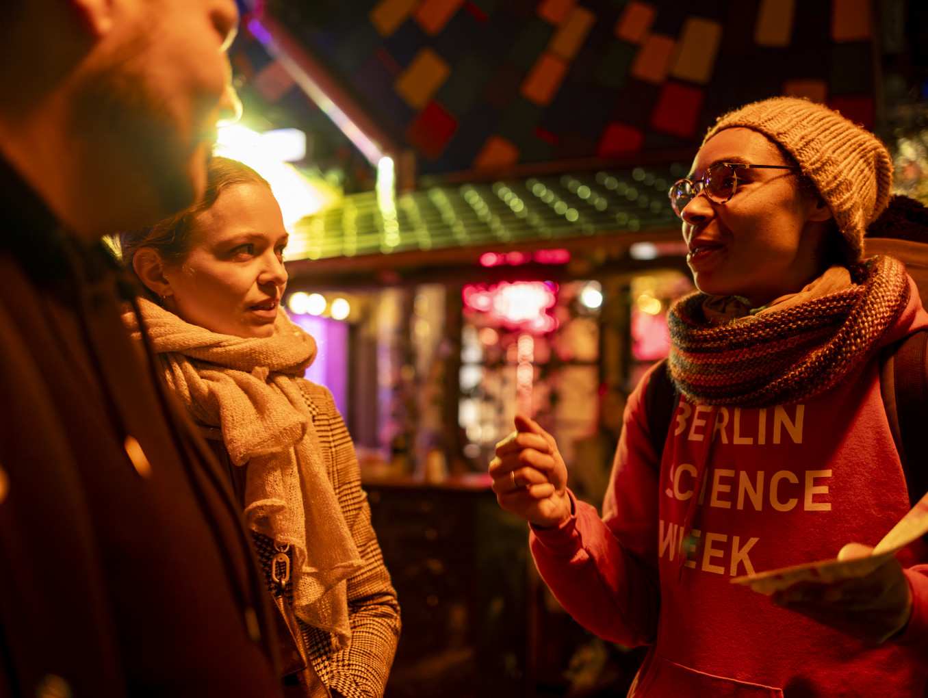 A woman wearing a red "Berlin Science Week" hoodie, a hat and a scarf, speaking to a couple