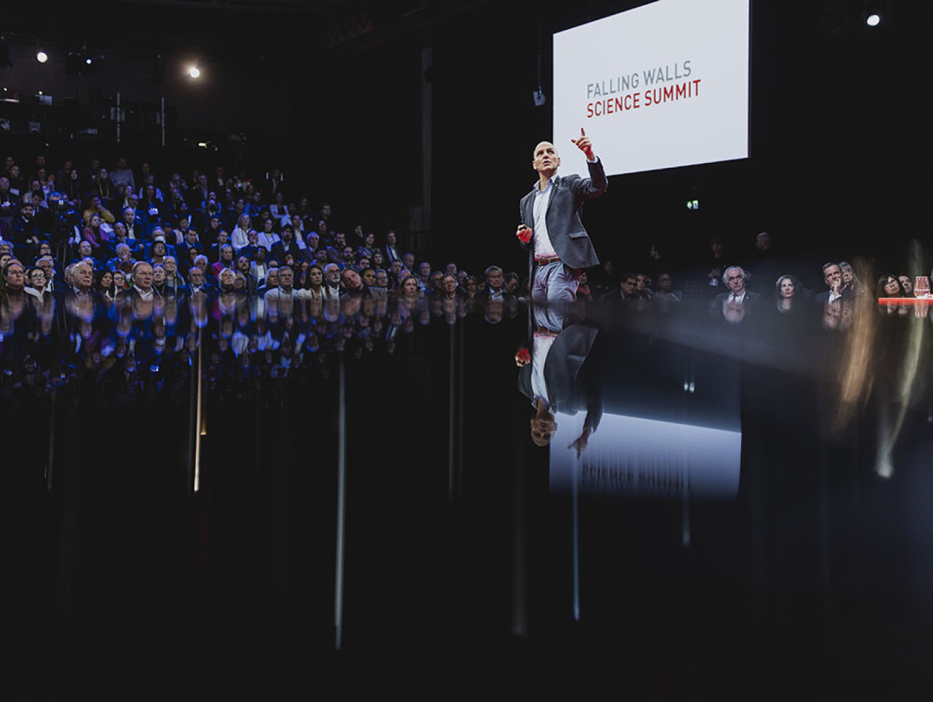 The image shows a speaker on stage at the Falling Walls Science Summit, addressing an audience seated in an auditorium. The speaker, dressed in a suit, is gesturing with one hand, emphasizing a point during their presentation. The stage has a reflective surface, creating a mirror-like effect that captures the speaker's image and the audience in front of them. In the background, a large screen displays the event name, "Falling Walls Science Summit." The lighting is dim, with focused spotlights on the speaker