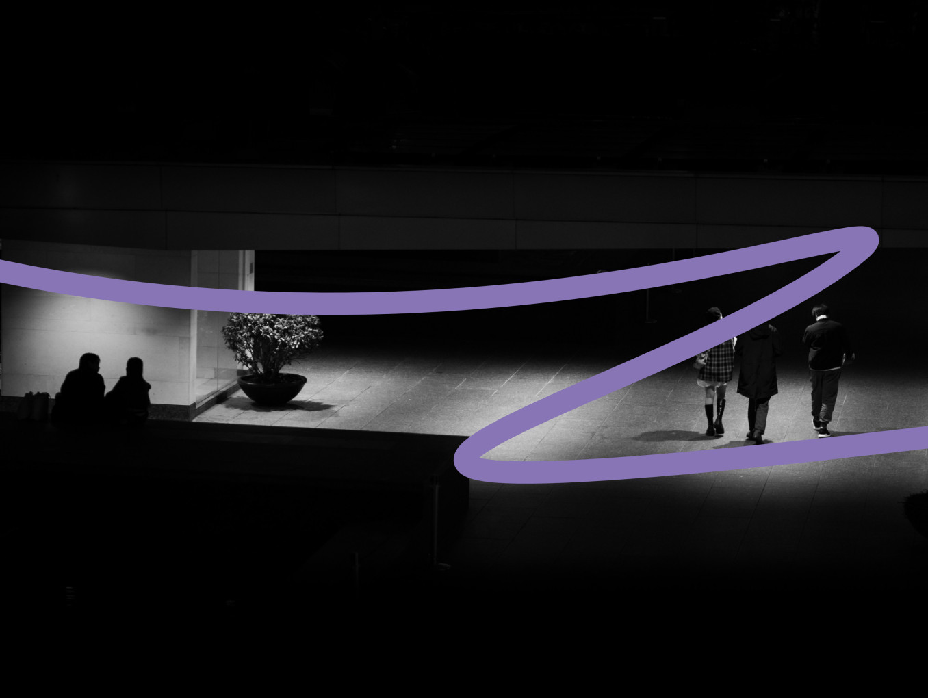 This image is showing a dimly lit urban scene at night. In the foreground, a wide walkway is illuminated by overhead lights. On the right side, three individuals walk away from the camera: two men and a woman, with the woman dressed in a checkered jacket or skirt, and the two men on either side of her. They walk towards a more shadowed area. To the left, under a bright light, two people sit on the floor, possibly talking, with a large potted plant next to them. The image has a minimalist, dark atmosphe