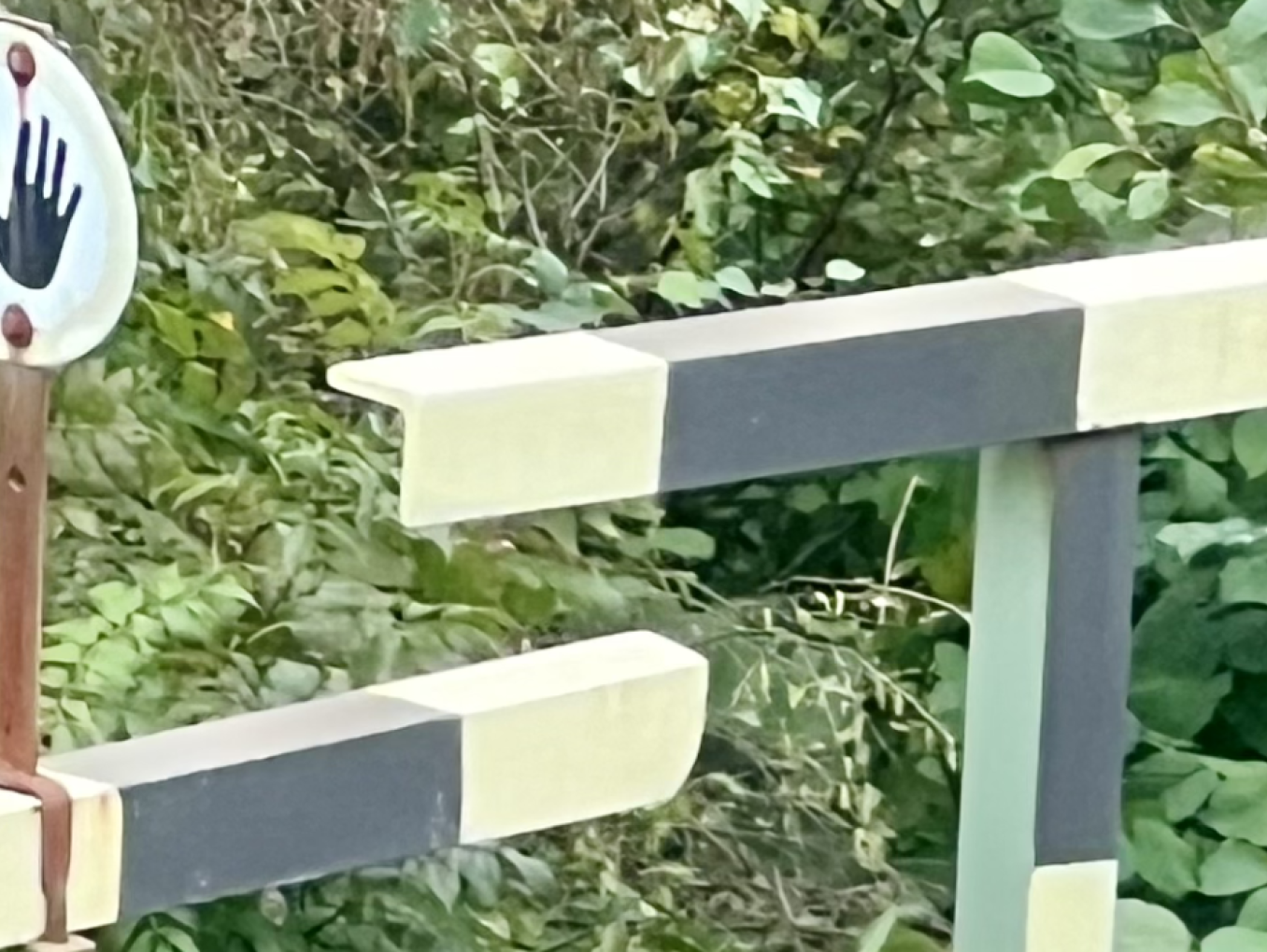The image shows a barrier gate, likely at a road or railway crossing, painted in alternating black and white stripes. Next to the gate is a circular sign with a handprint design, suggesting a stop or caution signal. The background is filled with dense green foliage, indicating a natural or rural setting. The design of the gate and the sign imply a request to halt, possibly to indicate restricted access or a need for caution at the crossing.