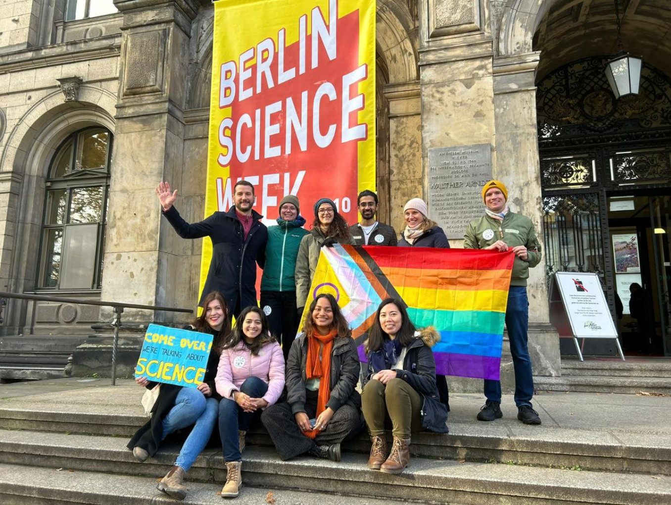 Soapbox Science Berlin and LGBTQ+ STEM Berlin teams pose with pride flag in front of Museum für Naturkunde.