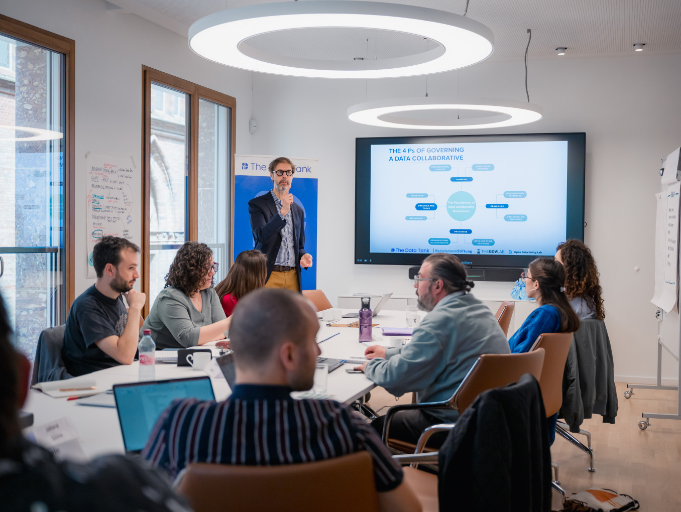 This image shows a meeting or workshop taking place in a modern conference room. Around a large table, several individuals are seated with laptops, notebooks, and coffee cups, actively listening to a speaker standing in front of a presentation screen. The speaker is holding a microphone and gesturing, explaining a slide titled "The 4 Ps of Governing a Data Collaborative." The slide contains a flowchart or diagram with various text bubbles, likely outlining the framework being discussed. 