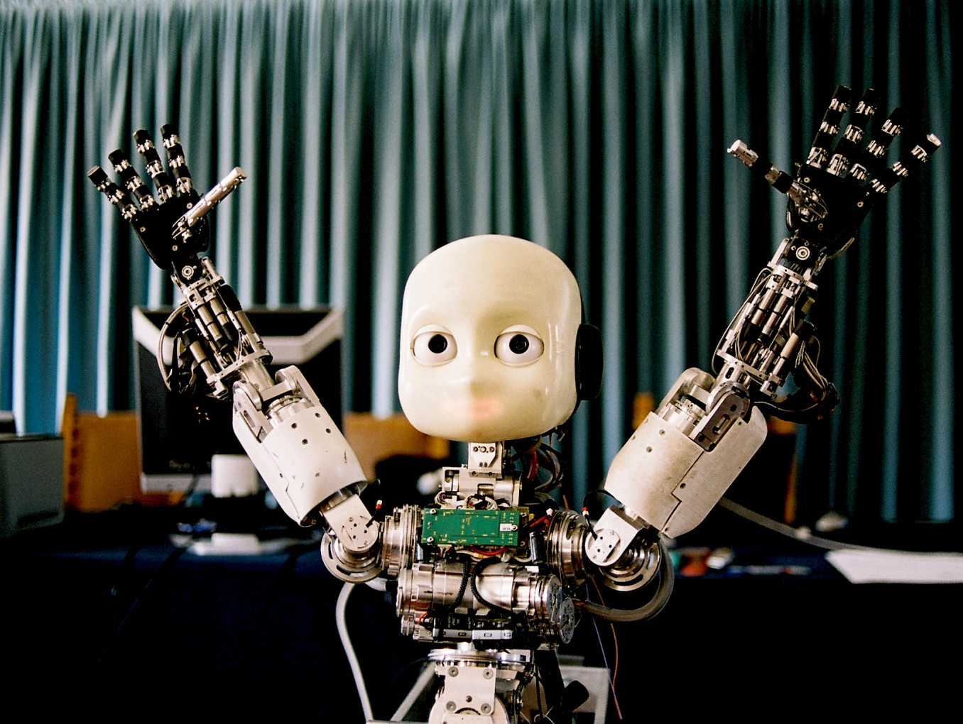Das Bild zeigt einen humanoiden Roboter mit einem minimalistischen Gesicht, großen Augen und erhobenen Armen. Der Roboter hat komplexe mechanische Hände mit vielen beweglichen Teilen und ist teilweise freigelegt, sodass seine inneren elektronischen Komponenten sichtbar sind. Im Hintergrund hängt ein Vorhang, und es befinden sich Computer und technische Ausrüstung auf einem Tisch.