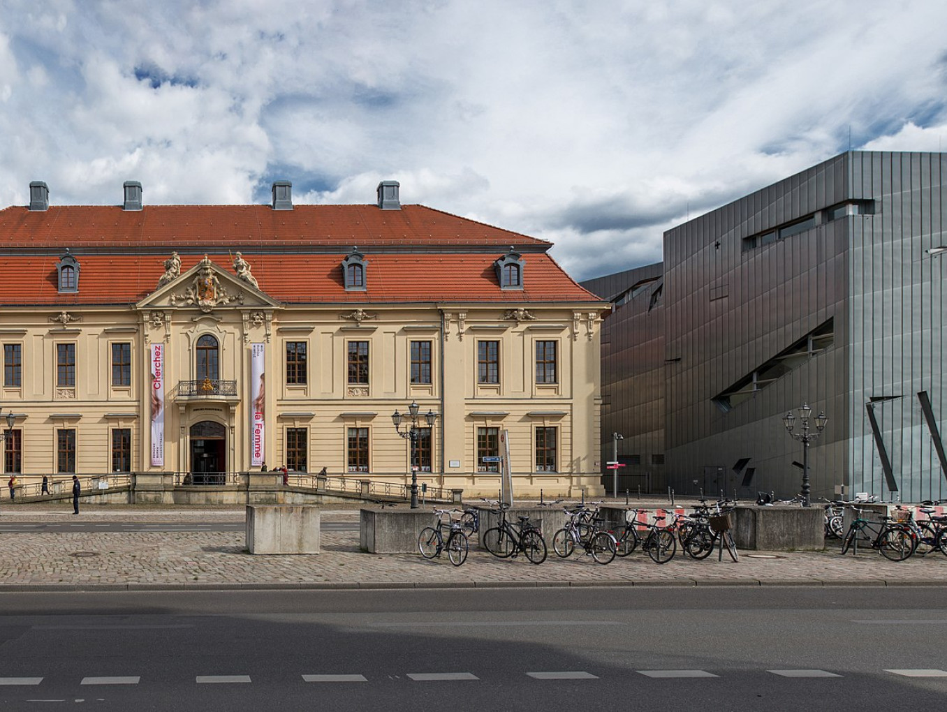 Two buildings: an old building on the left, a modern building on the right. In front there is a street. 
