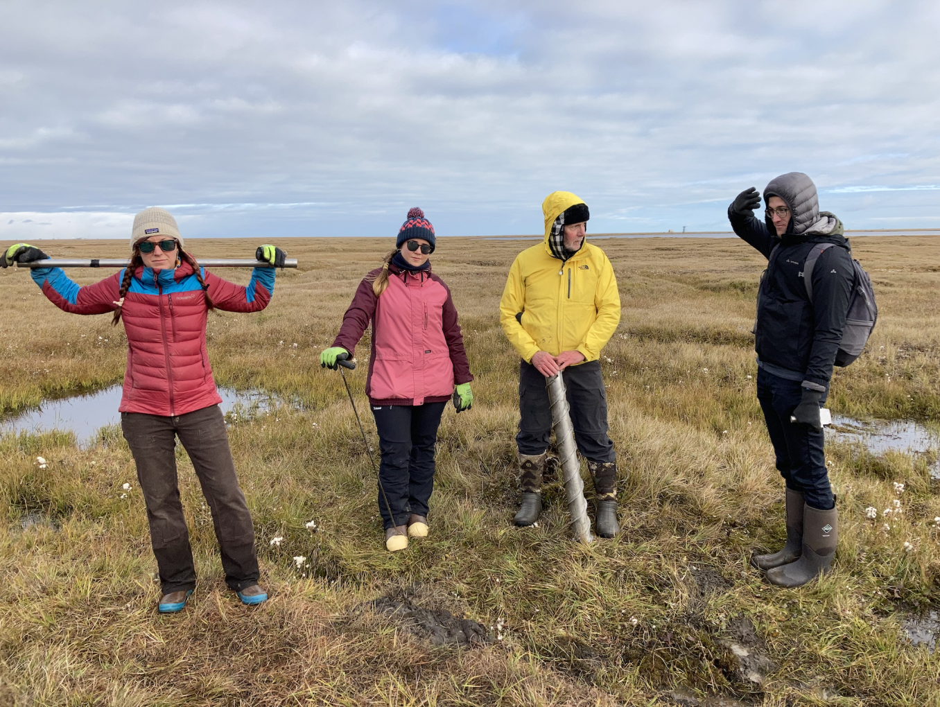 Two women and two men in bright-coloured outdoor clothing standing in the tundra with permafrost coring equipment.