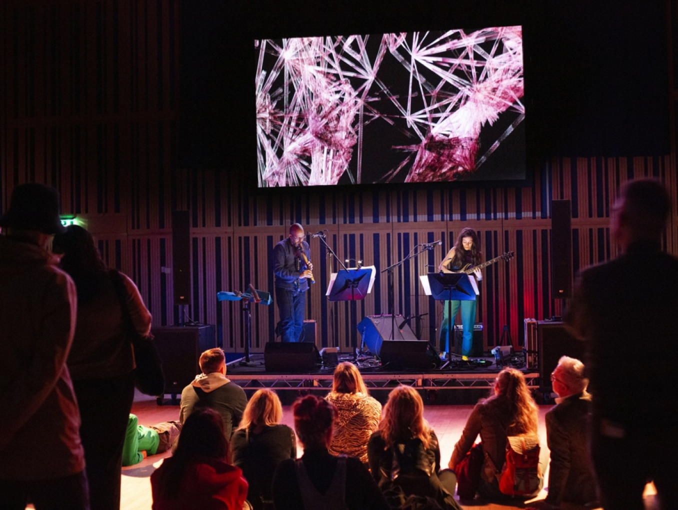 Two people on a stage playing instruments in front of a small crowd.