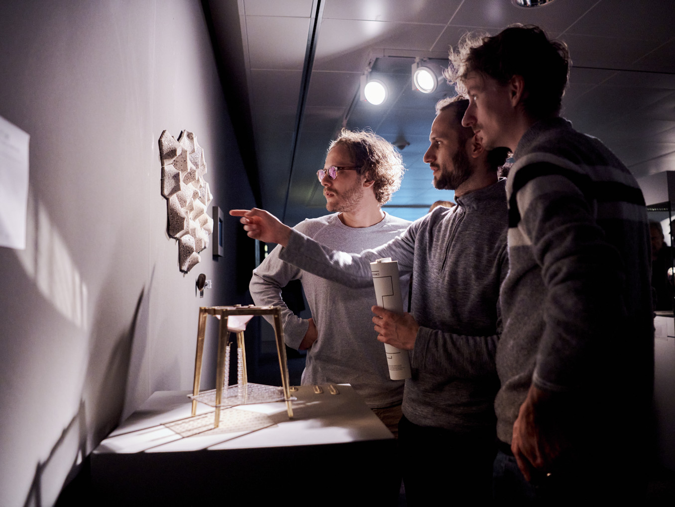 Three people in an exhibition pointing at an object on a wall. 