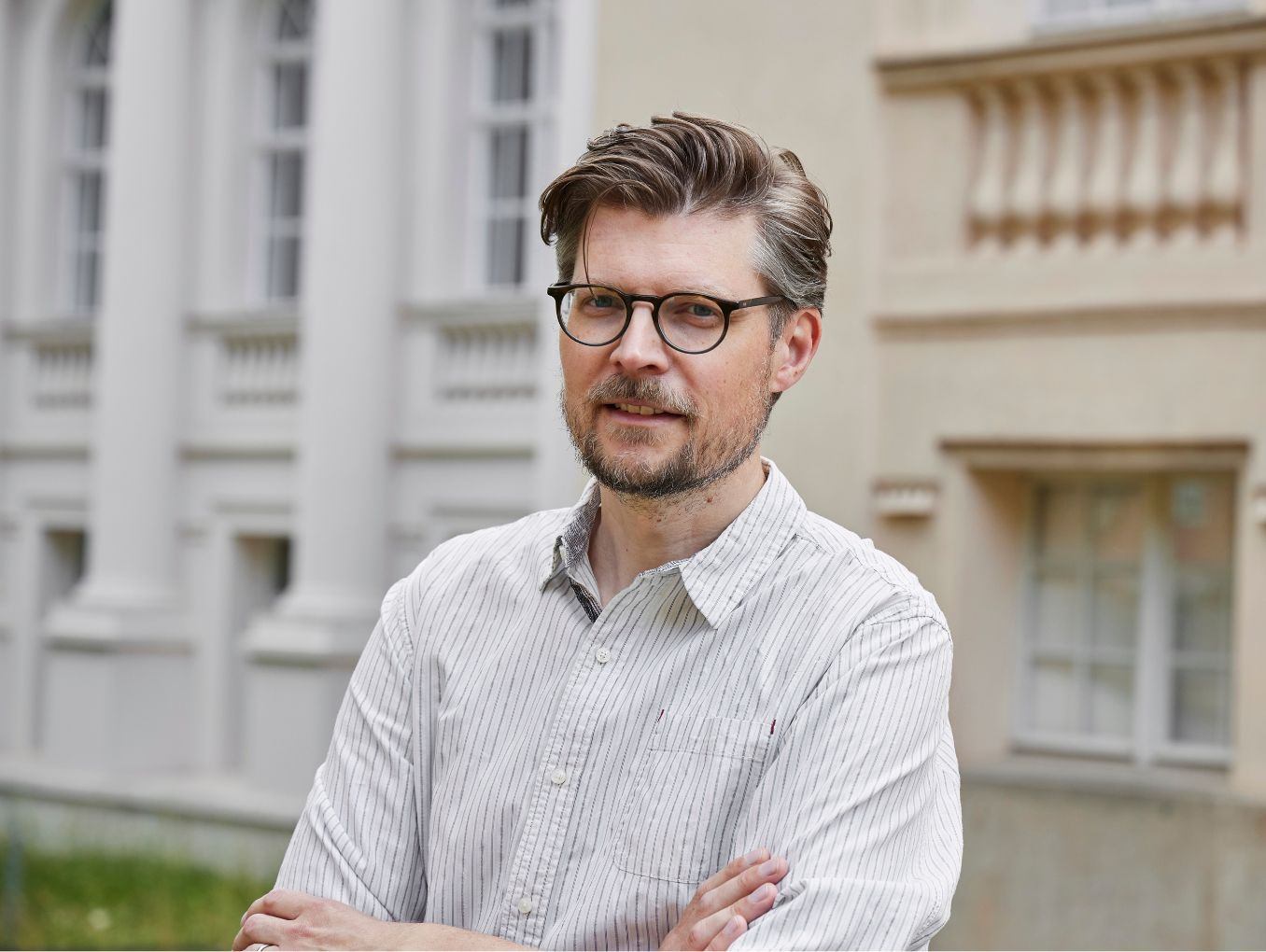 Tobias Krüger stands in a shirt and glasses with his arms crossed. 