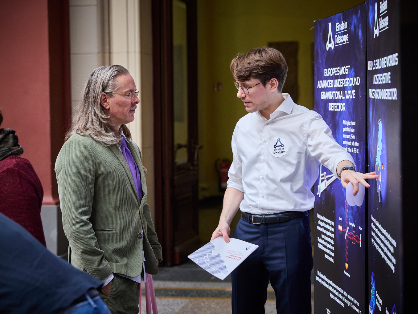 Two men stand at a board, one explains something to the other. 