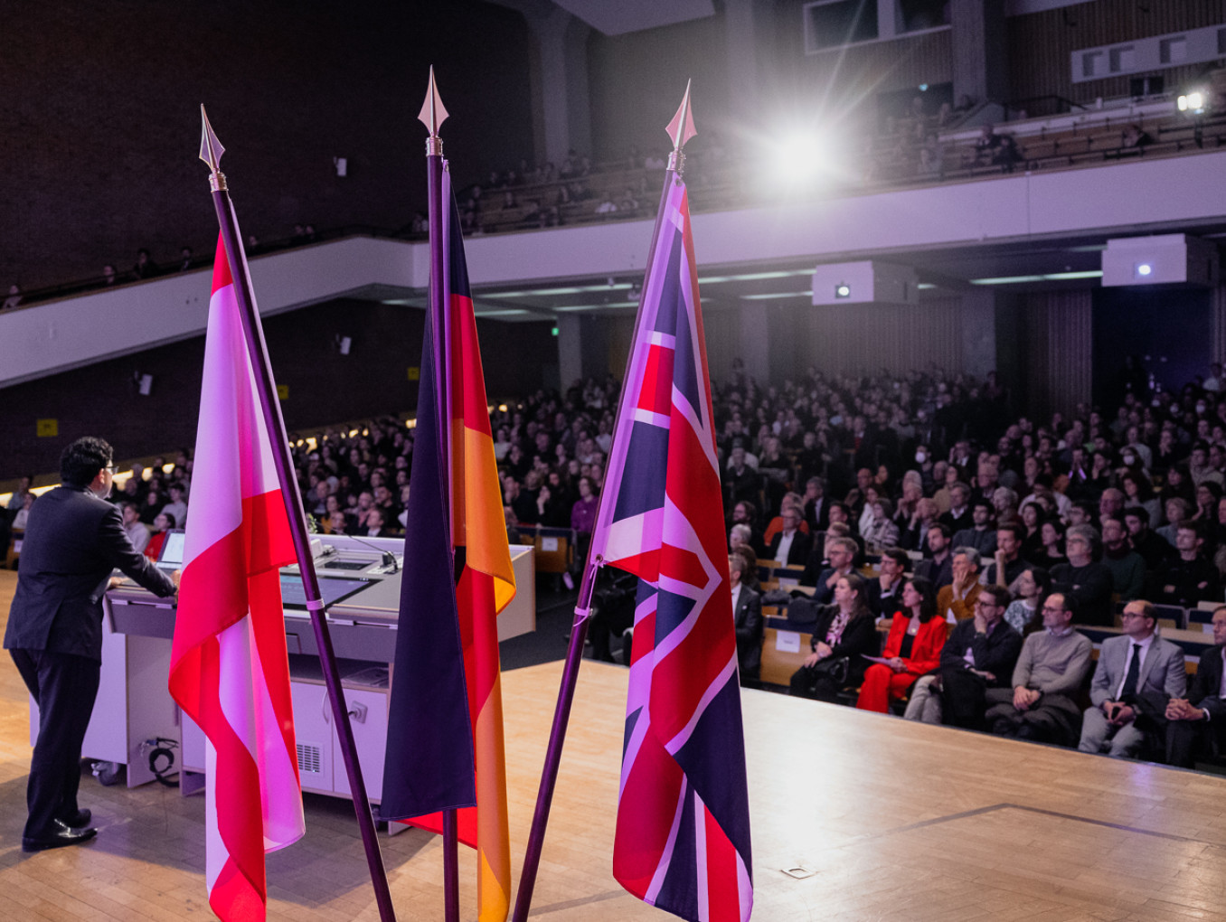 Lecture hall with many people, man at lectern, flags of the UK, Germany, Berlin