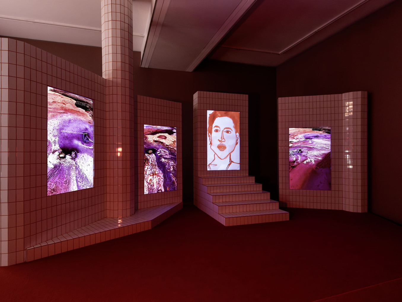 Four vertical screens in white tiles with red grout show videos of watercolor images and a portrait. 