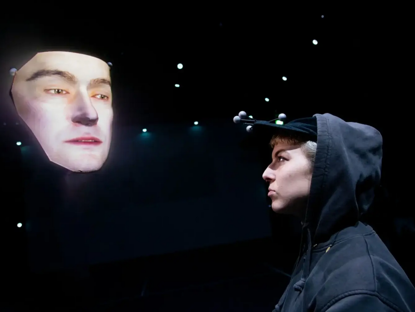 A person in a hoodie with motion-tracking sensors on their cap faces a large projected digital face in a dark room with small lights in the background.