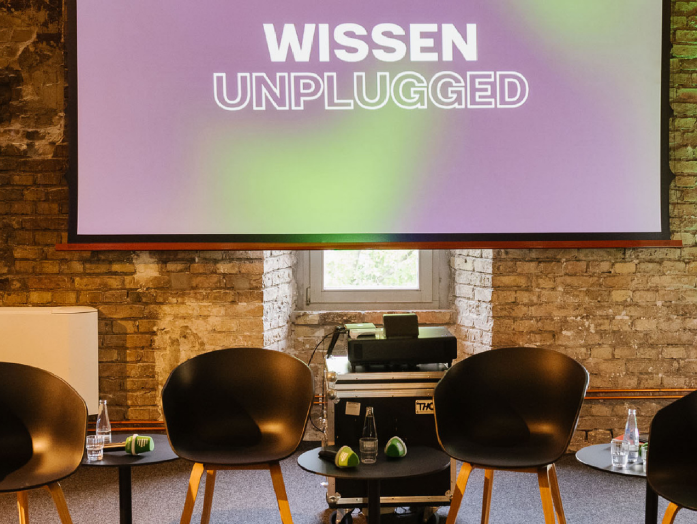 Fünf Stühle stehen vor einer Leinwand. Auf der Leinwand steht "Wissen Unplugged". Der Raum ist eine alte Ruine die renoviert worden ist als Veranstaltungslocation. 