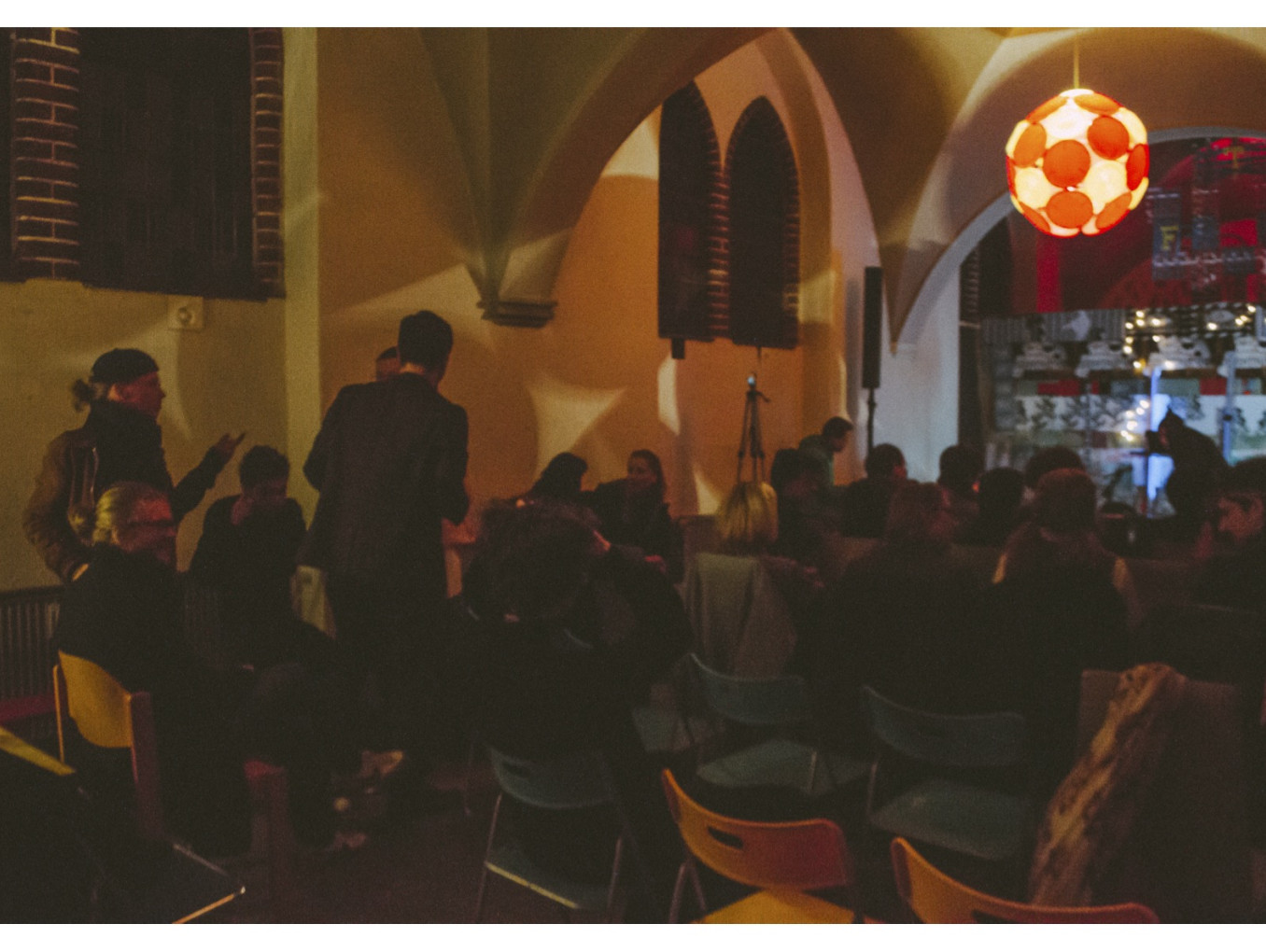A dimly lit indoor gathering with people seated on colorful chairs, facing a stage area with lights and decorations.