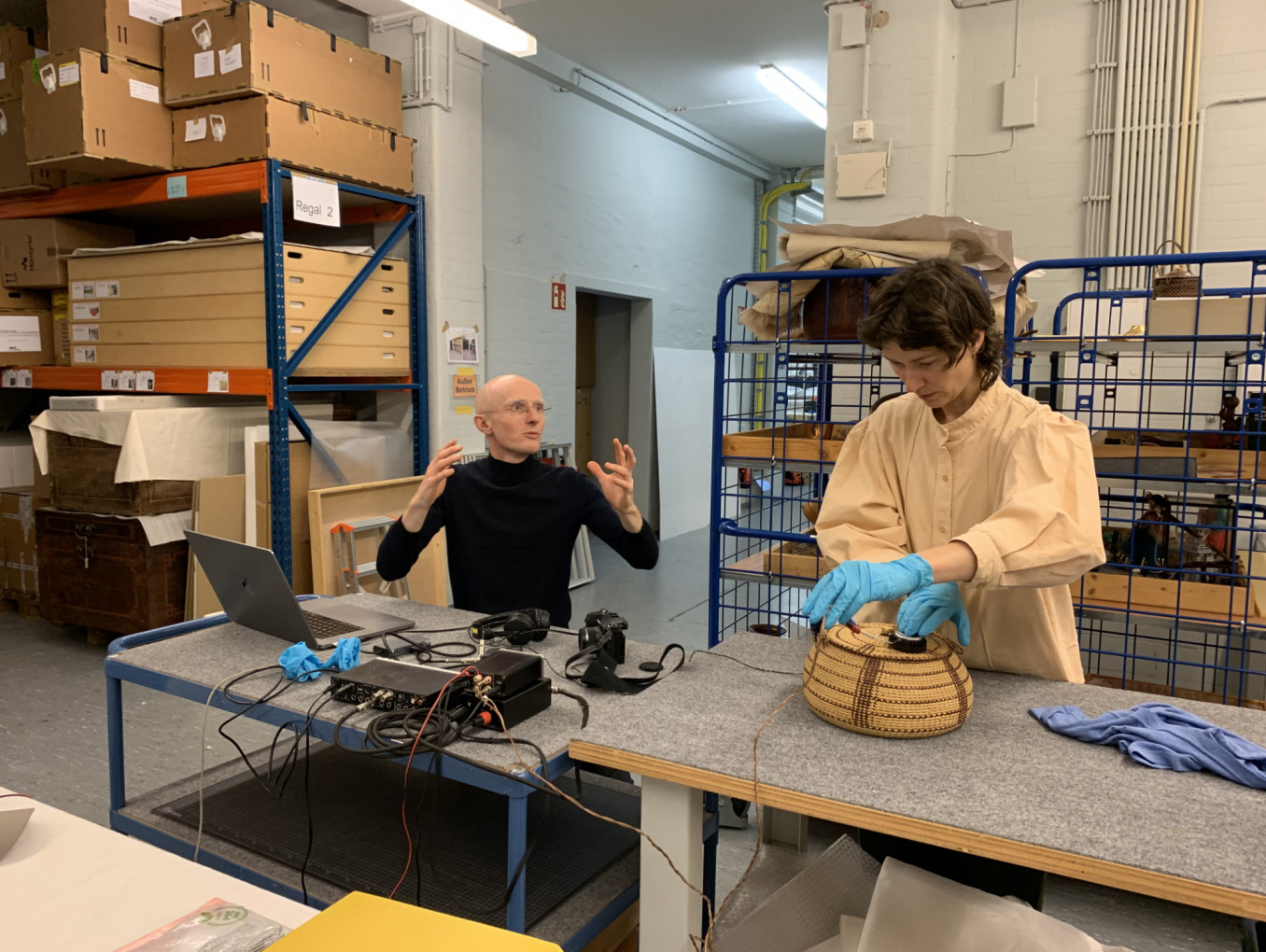 Two people are in a museum storage. One on the left side is seated and talking. One on the righ side standing and using a sound device over a museum collection item.