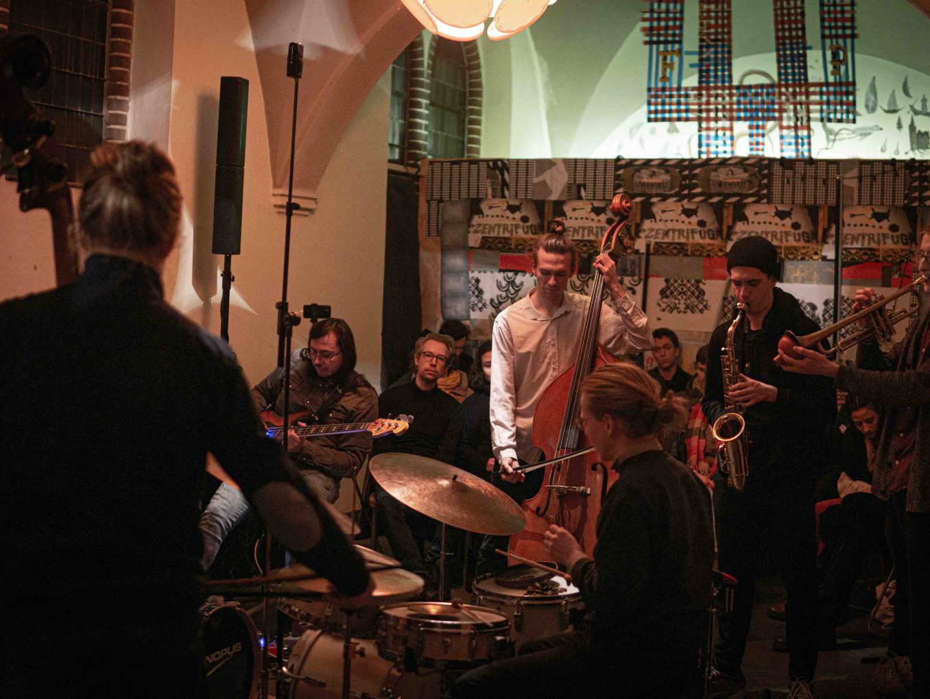 Eine kleine Jazzband spielt in einem gemütlichen, stimmungsvoll beleuchteten Raum vor einem aufmerksam lauschenden Publikum.
