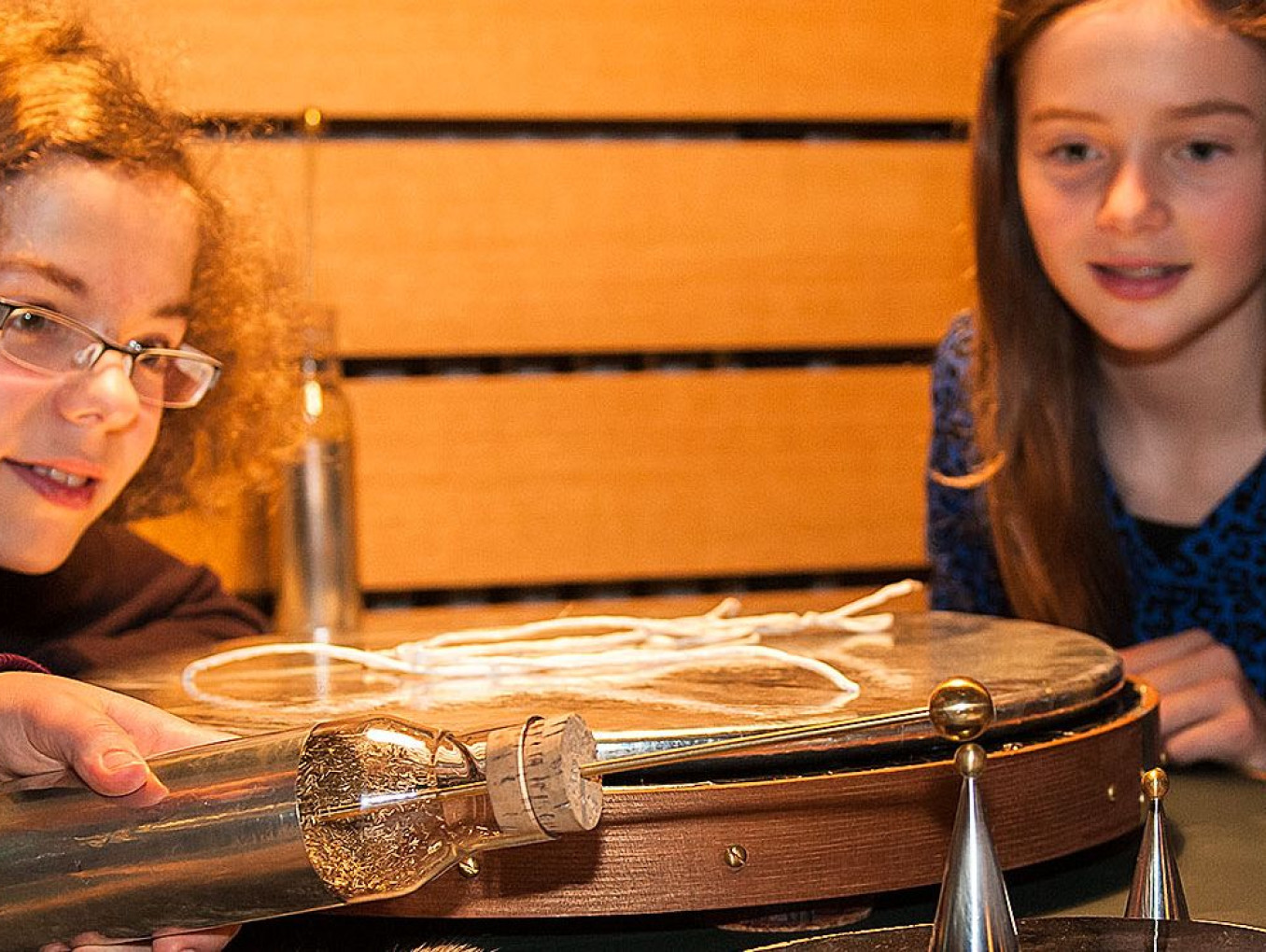 Fünf Mädchen experimentieren an einem Tisch. Ein Mädchen hält eine Glasflasche, aus der ein Stab an der Spitze ragt, an einen kleinen Metallkegel, der auf einer kleinen runden Platte mit glatter, schwarzer Oberfläche steht.