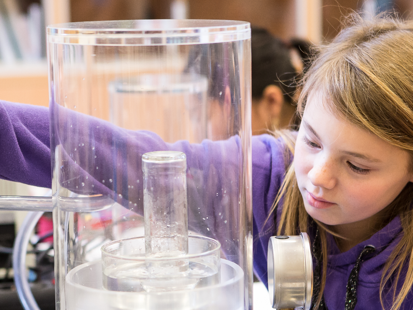 This image shows a young girl conducting a physics experiment with a vacuum bell jar at a laboratory table. It is a vacuum experiment in which the air is pumped out of a container.