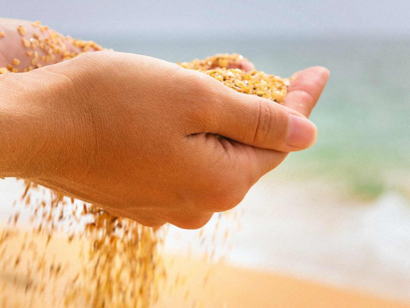 Zwei Hände halten eine Handvoll grobkörnigen Sand am Strand, während dieser langsam zwischen den Fingern hindurchrieselt. Im unscharfen Hintergrund sind das Meer und die sandige Küstenlinie bei hellem Tageslicht zu erkennen.