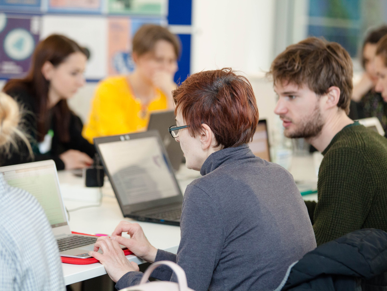 a group of different people in front of computers talking to each other and writing 
