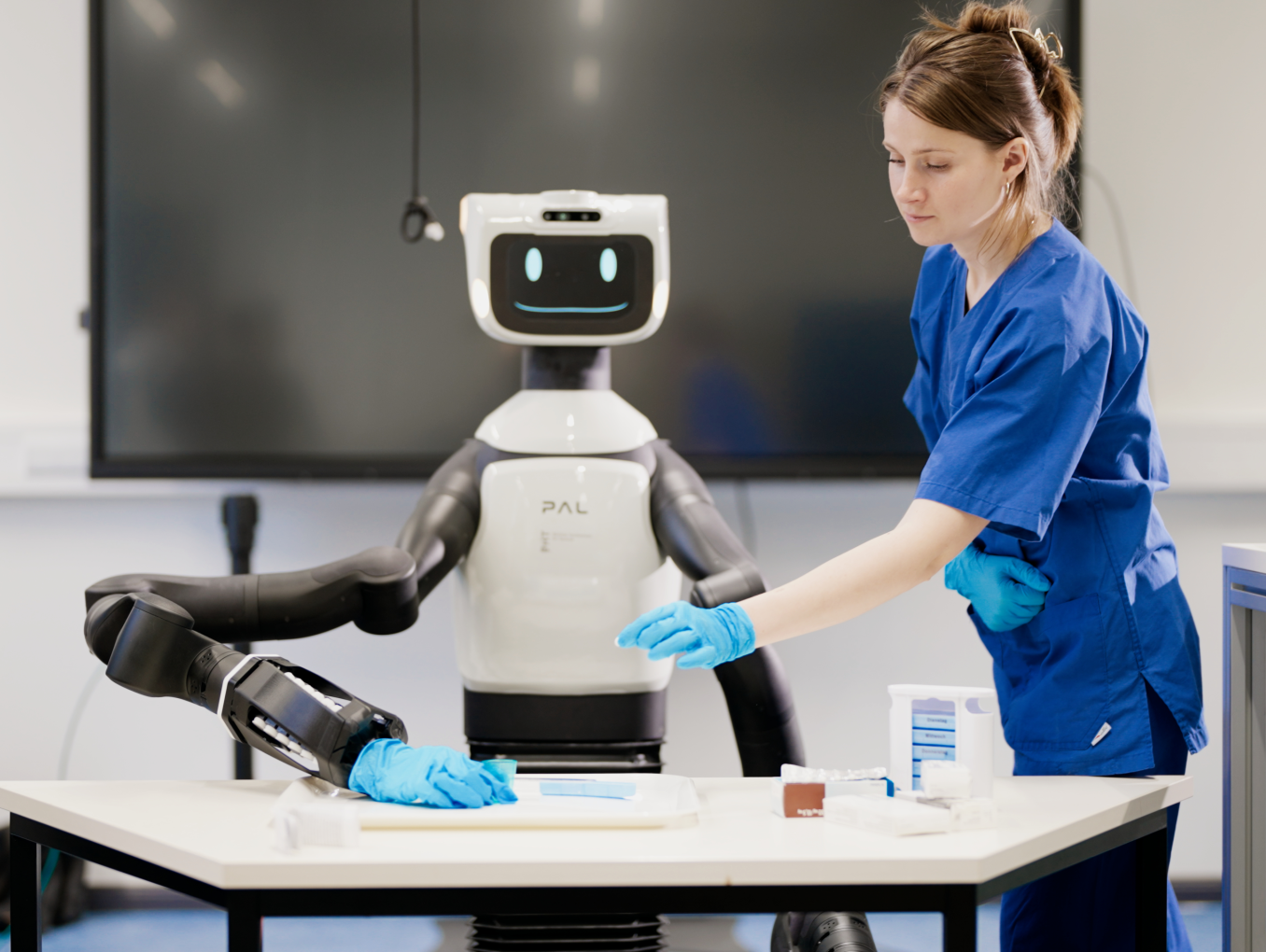 Der humanoide Roboter TIAGo Pro steht an einem Arbeitstisch mit Tabletten-Sortierschachteln, eine Person greift ebenfalls auf den Tisch zu.