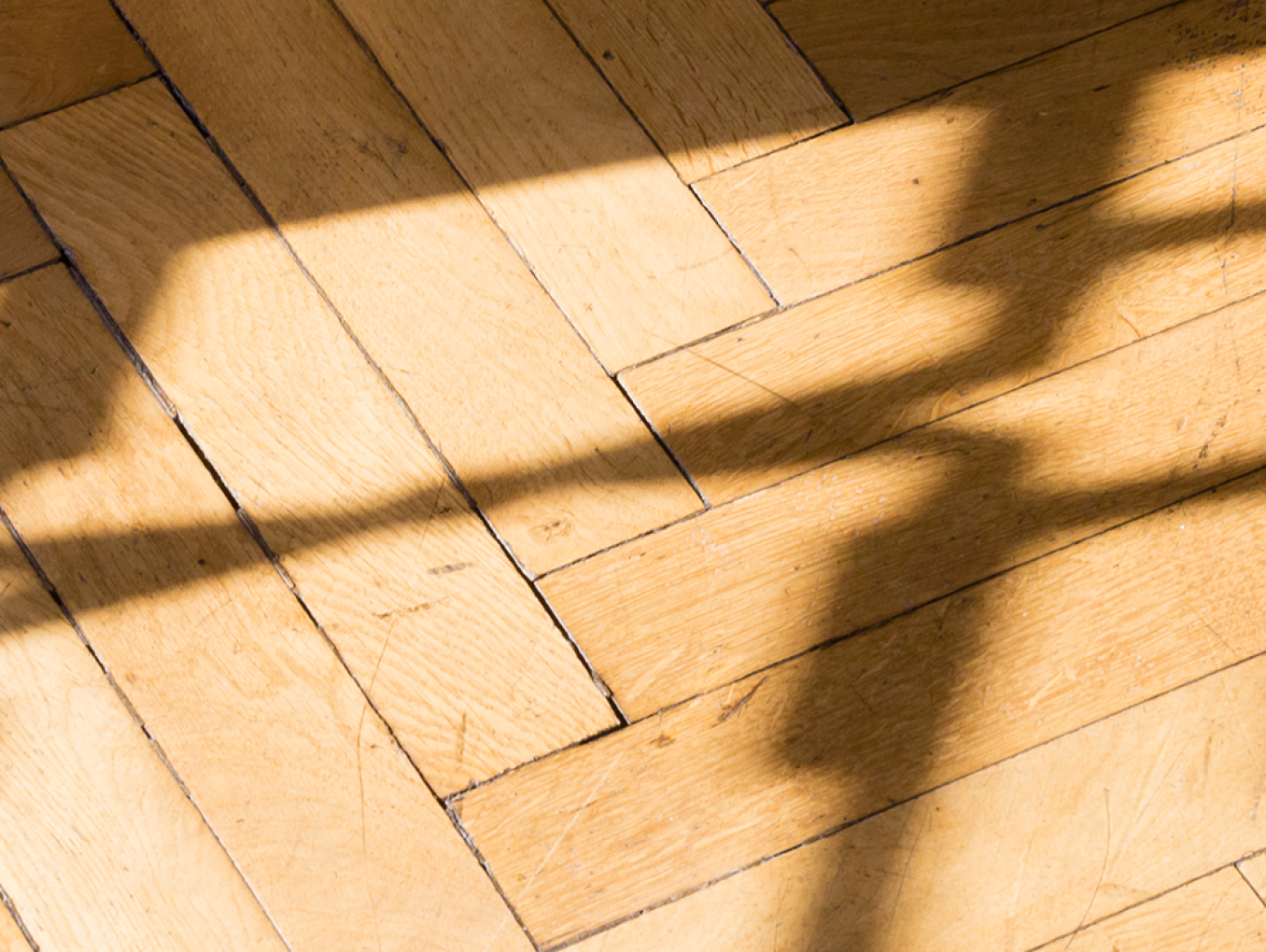 Nahaufnahme eines Parkettbodens aus hellem Holz im Fischgrätmuster. Über die Dielen fallen scharf gezeichnete Schatten einer schmiedeeisernen Balustrade, die durch Sonnenlicht abstrakte, geschwungene Linienmuster auf das Holz wirft.