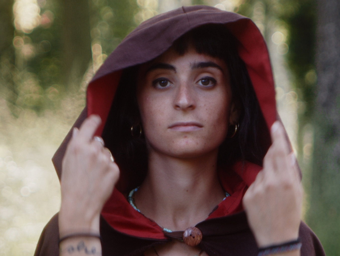 Head and shoulders of a woman in a red hooded cloak with out of focus forest in the background