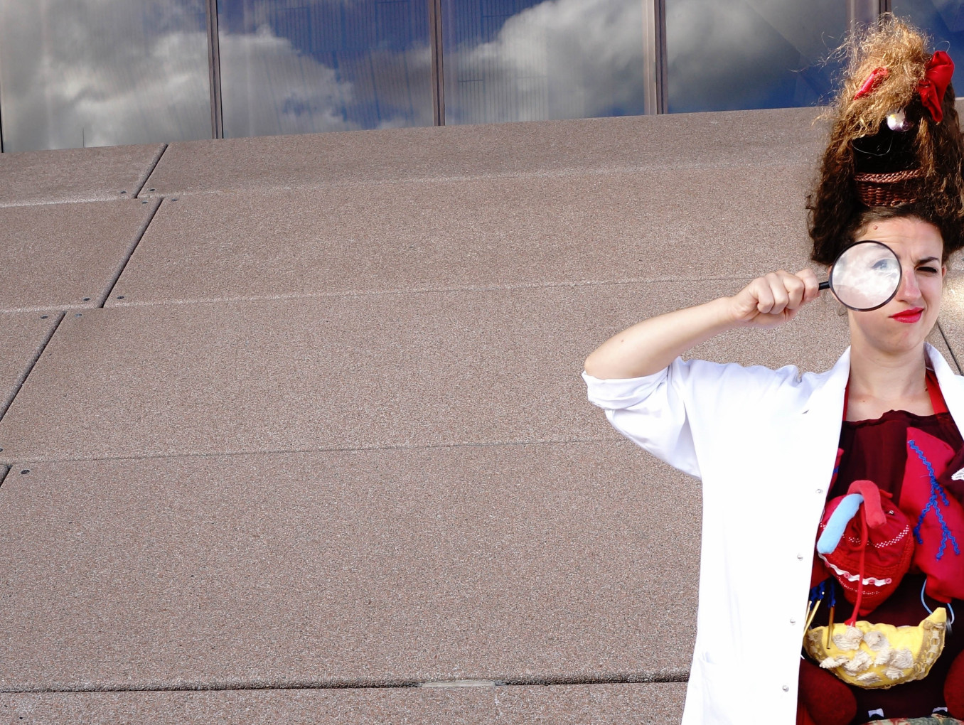 A woman with a tall hairstyle looks through a magnifying glass. She wears a labcoat that is open, revealing organs made from textiles.