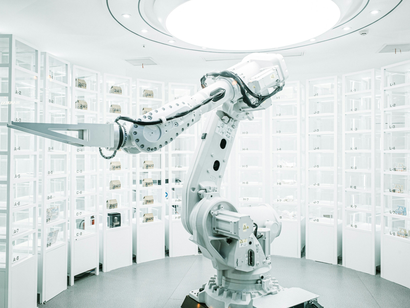 This photograph captures a robotic arm in a sterile, white-lit laboratory surrounded by tall shelving units filled with labeled containers. The scene reflects the use of advanced automation in precision manufacturing and pharmaceutical production environments.