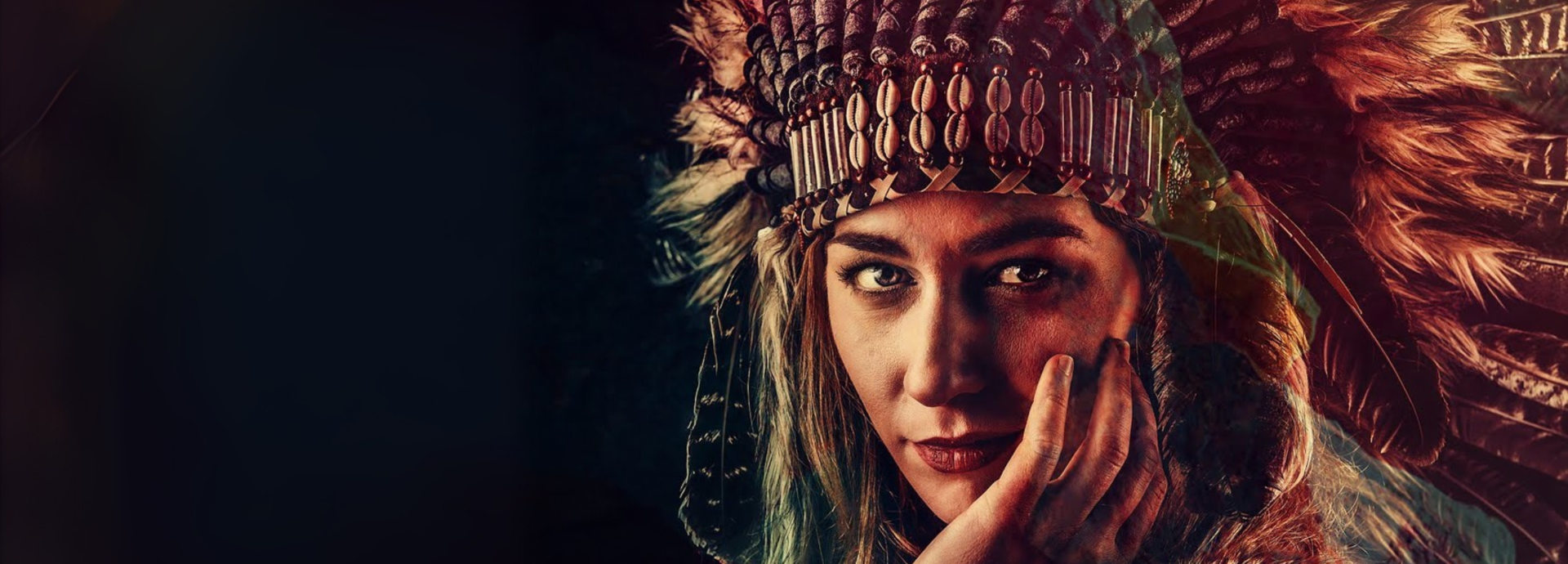 A woman wearing a traditional-style feathered headdress adorned with beads and shells, gazing directly at the camera with her hand resting against her face. The background is dark, highlighting her face and the detailed textures of the headdress.