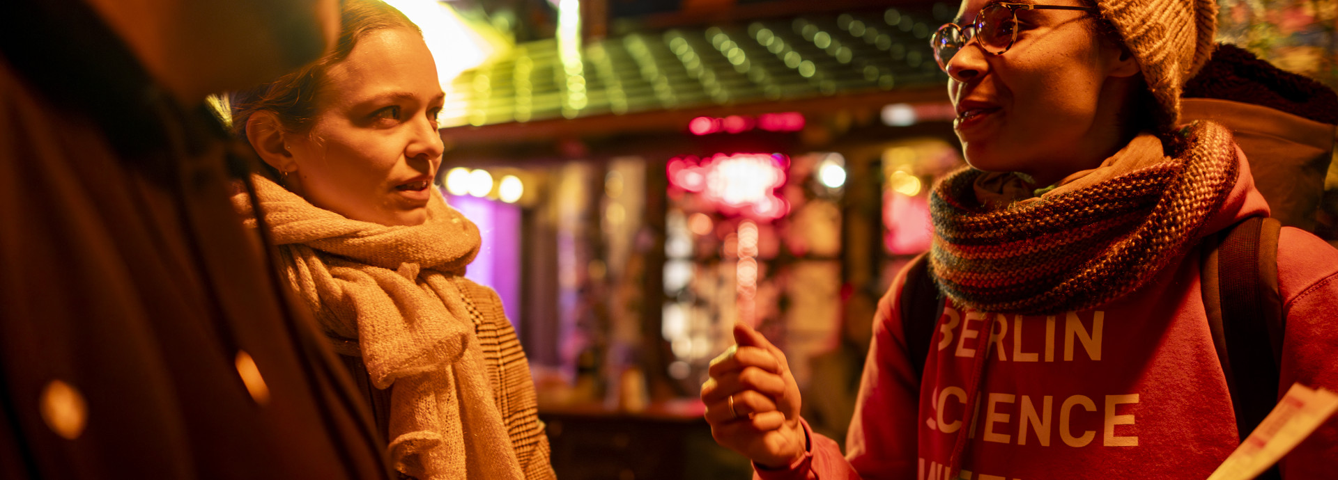 A woman wearing a red "Berlin Science Week" hoodie, a hat and a scarf, speaking to a couple