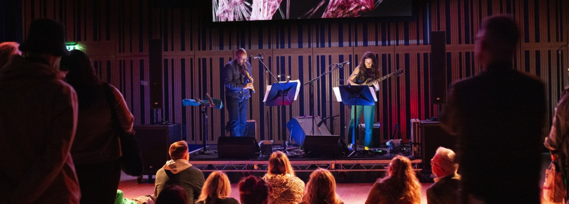 Two people on a stage playing instruments in front of a small crowd.