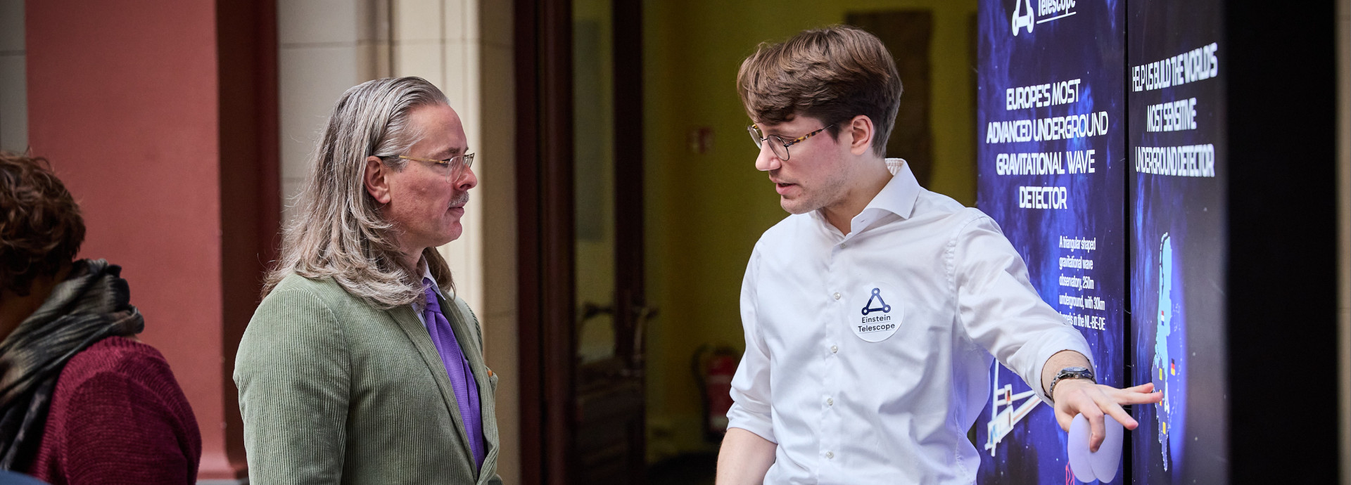 Two men stand at a board, one explains something to the other. 