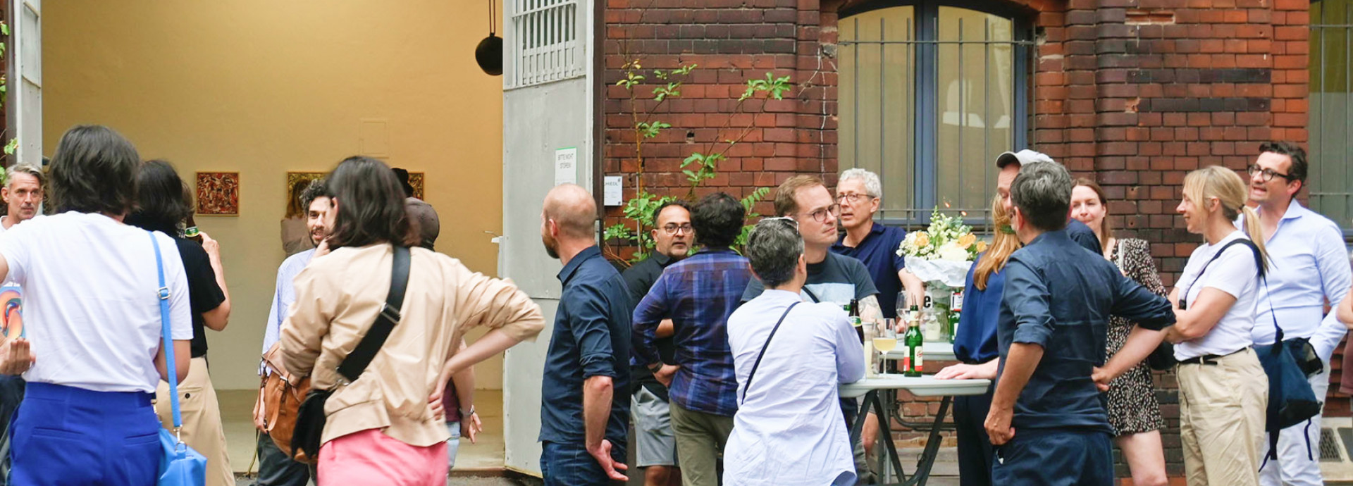 Eine Vernissage im Hinterhof der Urbanstraße 116 in Berlin, zahlreiche Menschen unterhalten sich in Gruppen, stehen mit Getränken oder sitzen an Tischen.