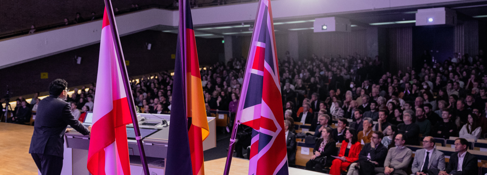 Lecture hall with many people, man at lectern, flags of the UK, Germany, Berlin