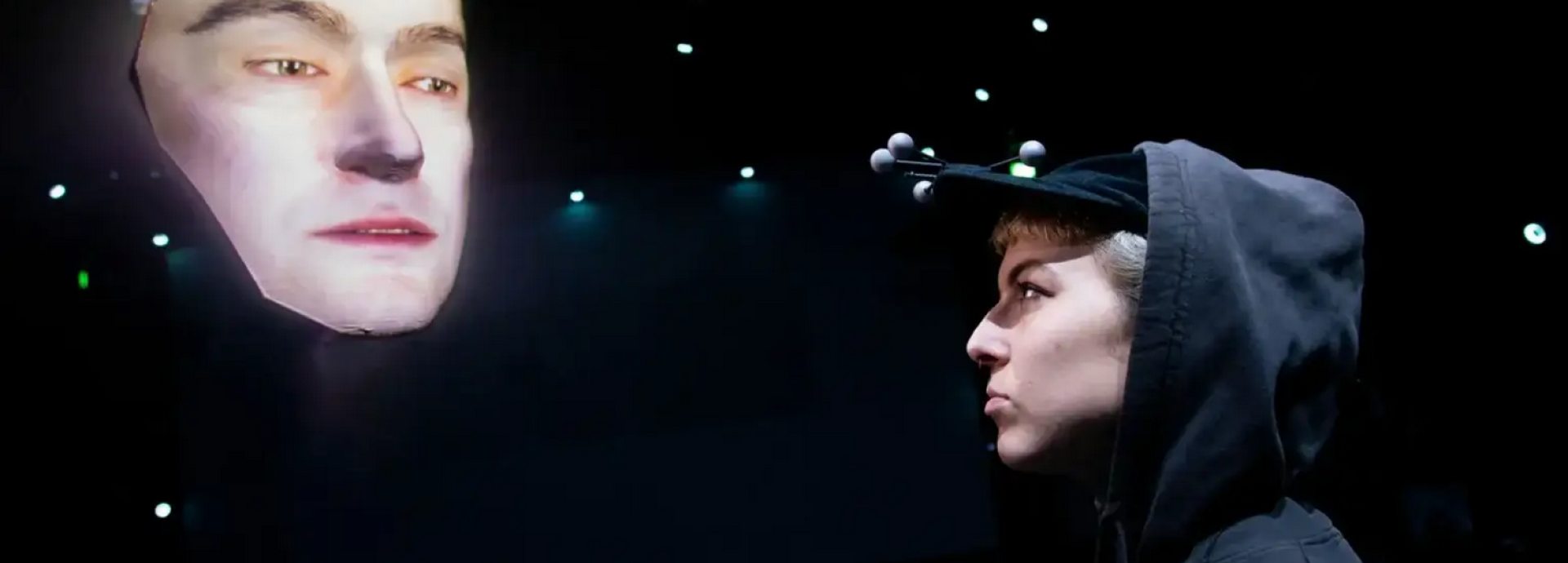 A person in a hoodie with motion-tracking sensors on their cap faces a large projected digital face in a dark room with small lights in the background.