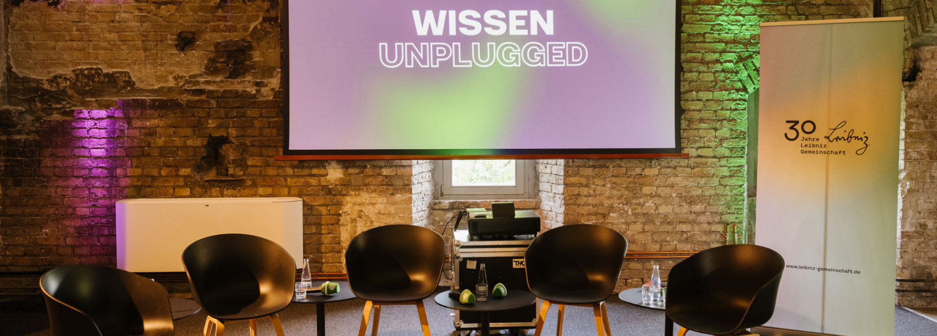 Fünf Stühle stehen vor einer Leinwand. Auf der Leinwand steht "Wissen Unplugged". Der Raum ist eine alte Ruine die renoviert worden ist als Veranstaltungslocation. 