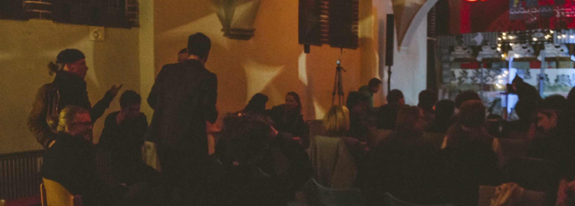 A dimly lit indoor gathering with people seated on colorful chairs, facing a stage area with lights and decorations.