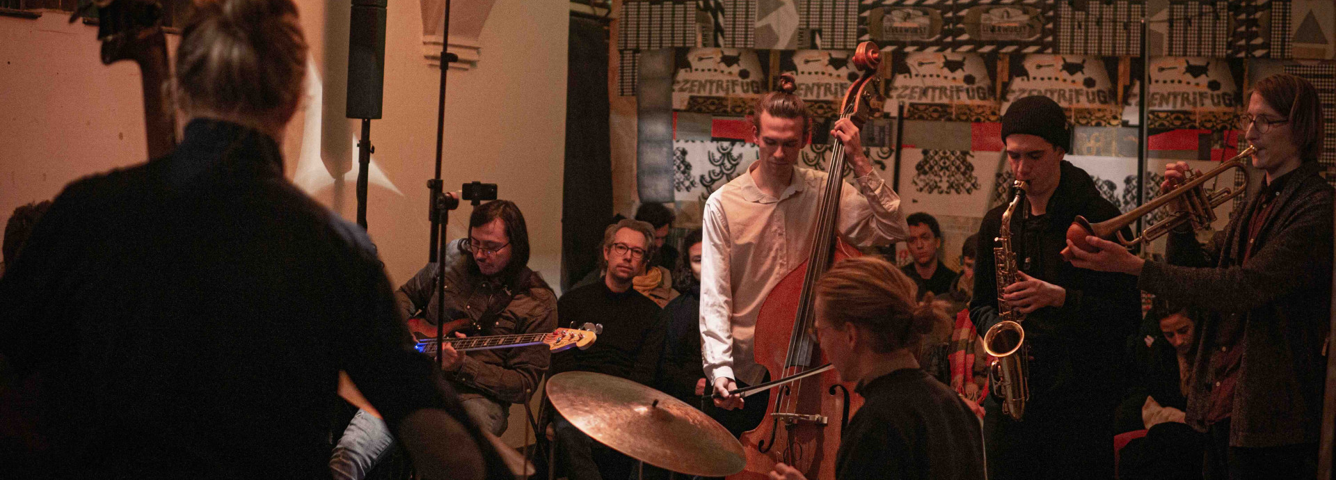Eine kleine Jazzband spielt in einem gemütlichen, stimmungsvoll beleuchteten Raum vor einem aufmerksam lauschenden Publikum.