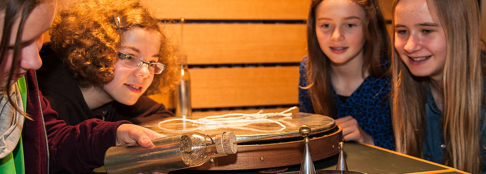 Fünf Mädchen experimentieren an einem Tisch. Ein Mädchen hält eine Glasflasche, aus der ein Stab an der Spitze ragt, an einen kleinen Metallkegel, der auf einer kleinen runden Platte mit glatter, schwarzer Oberfläche steht.