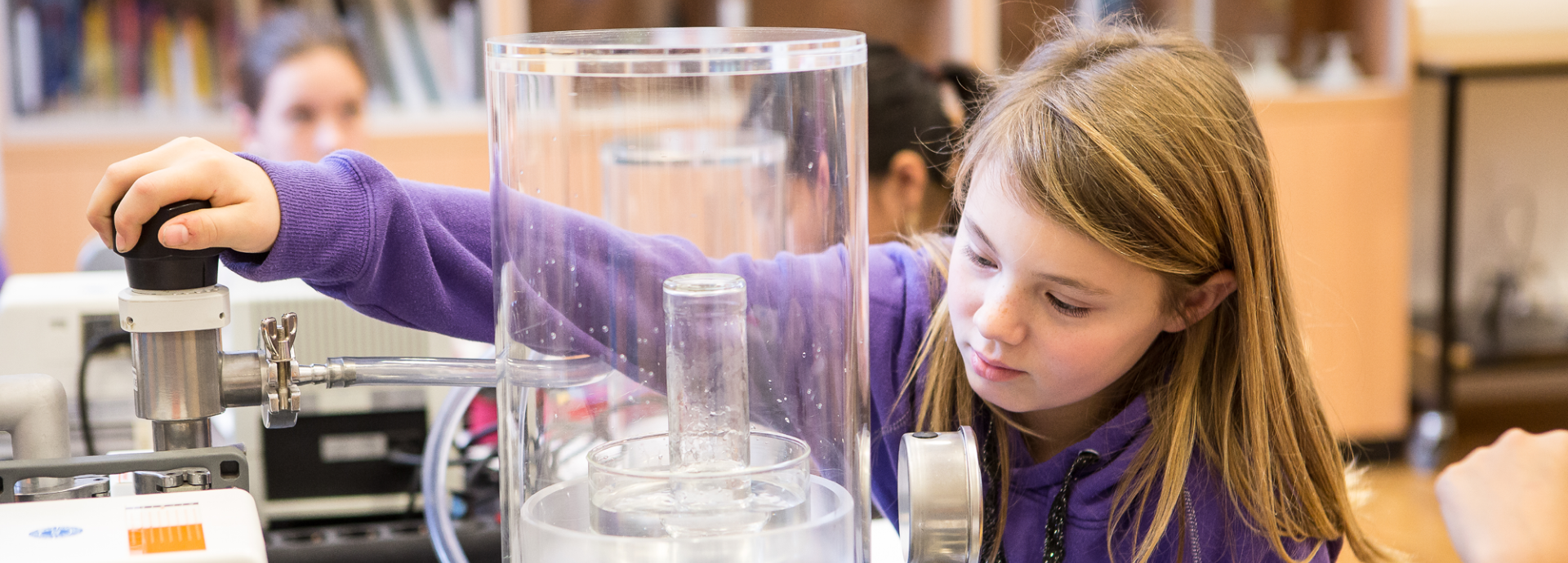 This image shows a young girl conducting a physics experiment with a vacuum bell jar at a laboratory table. It is a vacuum experiment in which the air is pumped out of a container.