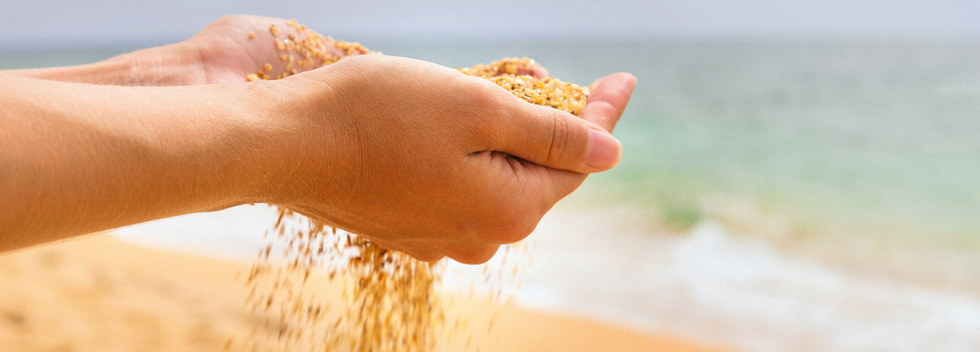 Zwei Hände halten eine Handvoll grobkörnigen Sand am Strand, während dieser langsam zwischen den Fingern hindurchrieselt. Im unscharfen Hintergrund sind das Meer und die sandige Küstenlinie bei hellem Tageslicht zu erkennen.