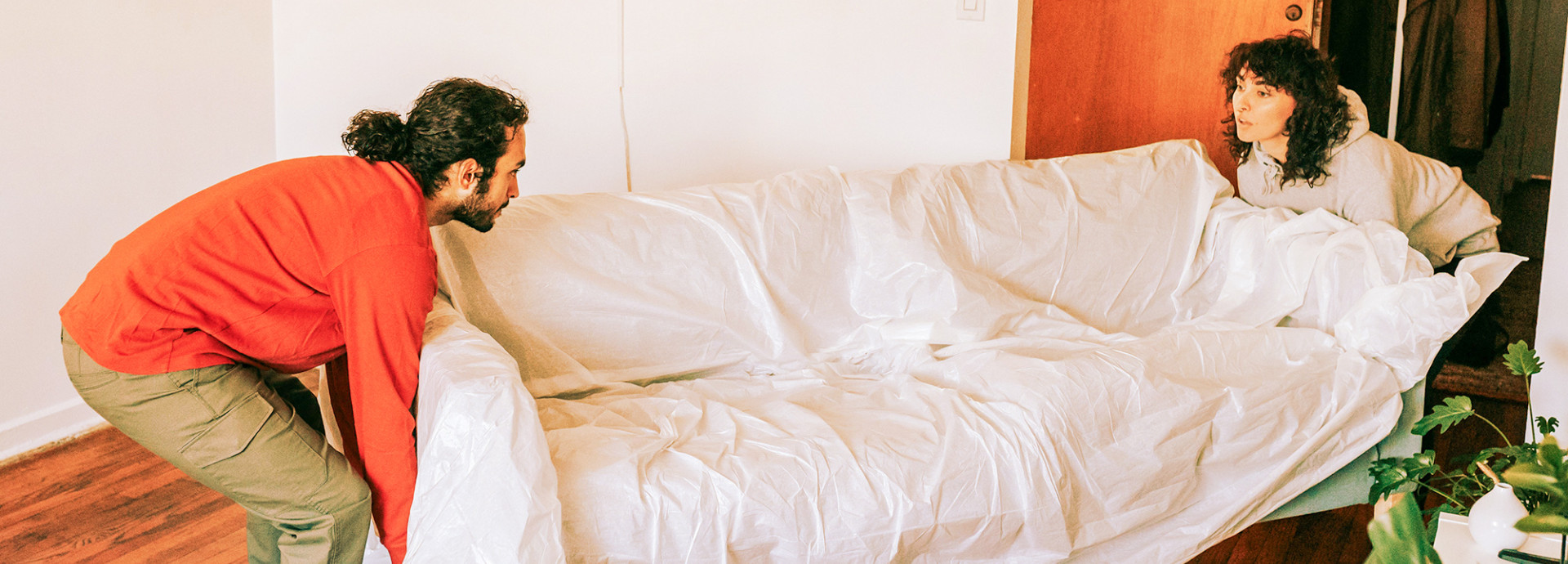 Horizontales Breitbild-Foto von zwei Personen, die ein mit weißer Plastikfolie abgedecktes Sofa in einem Raum bewegen. Links steht eine Person mit langen, lockigen, zu einem Dutt gebundenen Haaren, die einen leuchtend orangefarbenen Pullover und olivgrüne Cargohosen trägt und das Sofa von unten anhebt. Sie blickt konzentriert nach rechts. Rechts steht eine zweite Person mit kurzen, dunklen, lockigen Haaren und einem grauen Kapuzenpullover, die das Sofa von der anderen Seite anhebt und nach links blickt. Der