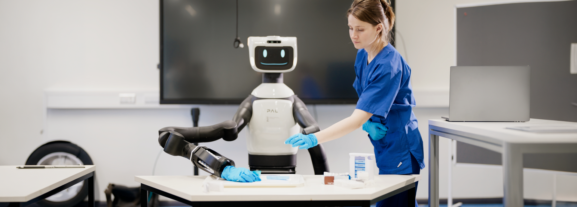Der humanoide Roboter TIAGo Pro steht an einem Arbeitstisch mit Tabletten-Sortierschachteln, eine Person greift ebenfalls auf den Tisch.