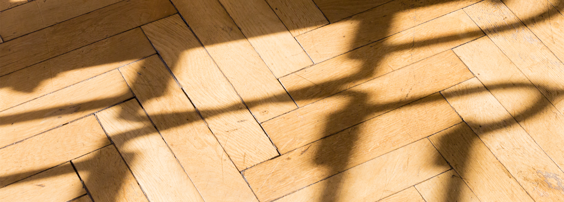 Nahaufnahme eines Parkettbodens aus hellem Holz im Fischgrätmuster. Über die Dielen fallen scharf gezeichnete Schatten einer schmiedeeisernen Balustrade, die durch Sonnenlicht abstrakte, geschwungene Linienmuster auf das Holz wirft.