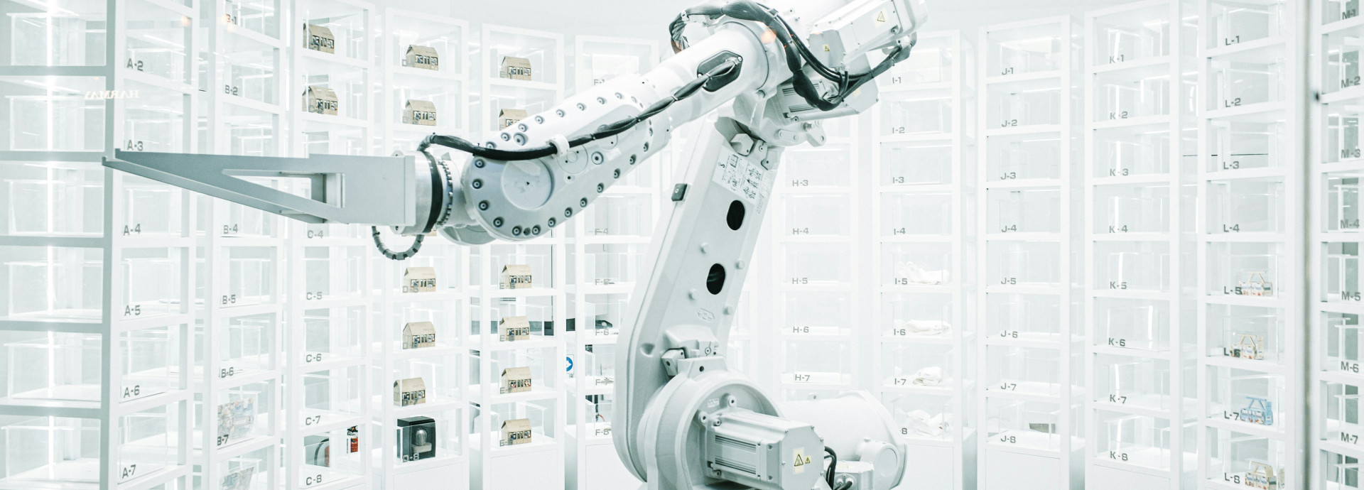 This photograph captures a robotic arm in a sterile, white-lit laboratory surrounded by tall shelving units filled with labeled containers. The scene reflects the use of advanced automation in precision manufacturing and pharmaceutical production environments.