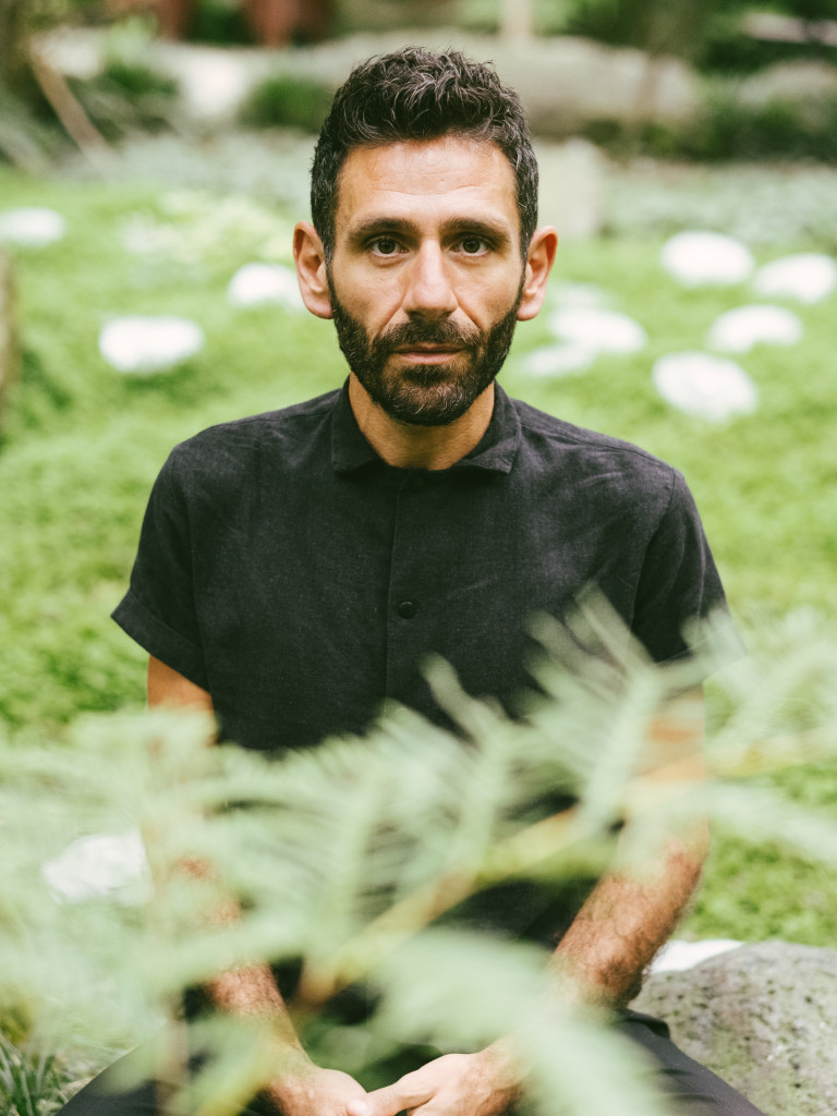 This image depicts a man sitting outdoors, with lush greenery in the background. The man is seated on a rock, facing the camera, with a calm and contemplative expression. He has short, curly dark hair and a neatly trimmed beard. He is wearing a simple, dark-colored button-up shirt, and his hands are gently clasped in his lap. In the foreground, some plants are slightly out of focus, adding depth to the composition. The natural setting gives a peaceful and serene atmosphere to the portrait.